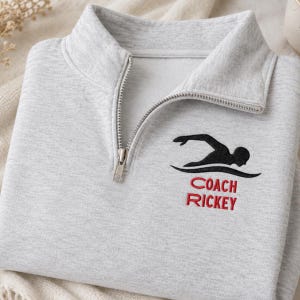 May include: A light gray quarter-zip sweatshirt featuring a silver metal zipper and a black embroidered graphic of a person swimming. Below the swimmer, the text COACH RICKEY is embroidered in red capital letters on the left chest area.
