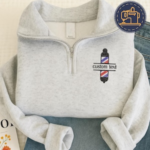 May include: A light gray quarter-zip sweatshirt with a barber pole design and the words "custom text". The barber pole is black, red, and blue. The sweatshirt is folded on top of blue jeans.