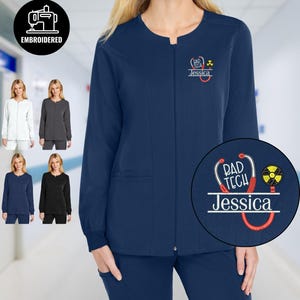 May include: Navy blue medical scrub jacket with a zip fastening and embroidered details. The jacket has a custom design with the text "RAD TECH" and a name. Additional colours shown: white, grey, and black.