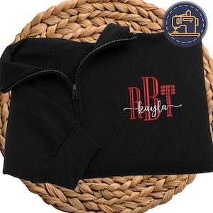 May include: Black quarter-zip sweatshirt with embroidered red initials and the name "kayla" in a cursive font. The sweatshirt has a black zipper and is folded on a woven, circular surface. A sewing machine patch is in the upper right corner.