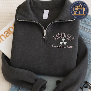 May include: Dark gray quarter-zip sweatshirt with the word "RADIOLOGY" embroidered in pink, along with a radiation symbol and the name "Kristin, LMRT". The sweatshirt has a silver zipper and a small tag.