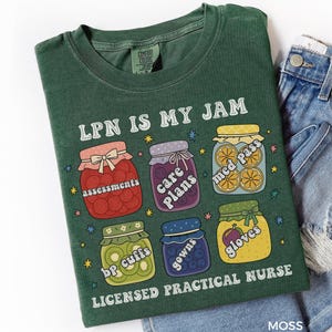 May include: Moss green t-shirt with the text "LPN IS MY JAM" and illustrations of jam jars containing medical-related items. The shirt also features the words "LICENSED PRACTICAL NURSE". The shirt is paired with denim shorts.