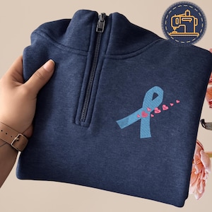 May include: A navy blue quarter-zip sweatshirt featuring a light blue ribbon with pink heart accents. The sweatshirt has a zipper closure and a small embroidered sewing machine patch. The image is of a sweatshirt.