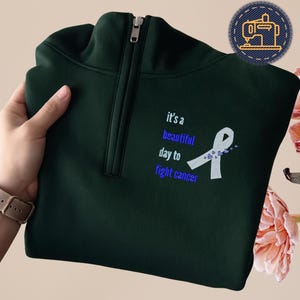 Puede incluir: Sudadera verde oscuro con cremallera de cuarto de círculo y un diseño de cinta blanca y azul. El texto "it's a beautiful day to fight cancer" está bordado en la parte delantera. Cremallera plateada.