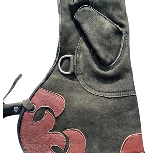 May include: A dark green leather glove with a burgundy accent design. The glove features a silver metal ring and a strap. The burgundy design is a stylized flame shape. The glove is designed for protection.