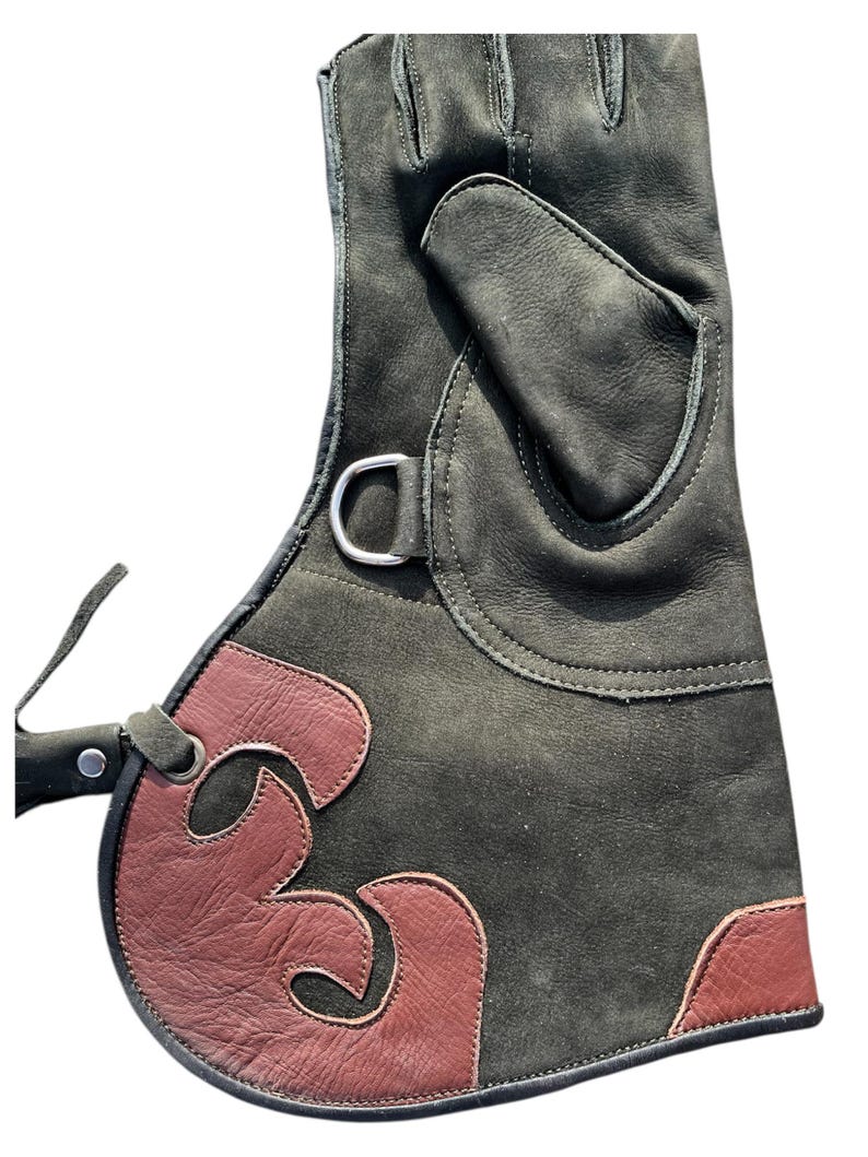 May include: A dark green leather glove with a burgundy accent design. The glove features a silver metal ring and a strap. The burgundy design is a stylized flame shape. The glove is designed for protection.
