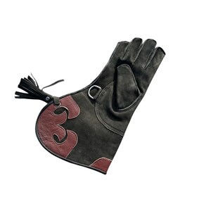 Op de afbeelding: Een donkergroene suède valkeniershandschoen met bruine leren details. De handschoen heeft een metalen ring, een decoratief bruin leren ontwerp en een kwastje. Ontworpen om de hand te beschermen tijdens de valkerij.