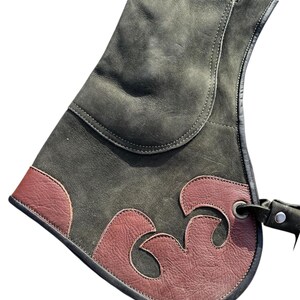 May include: A dark green leather falconry glove with a decorative brown leather accent. The glove features a curved design and a strap with a metal eyelet for securing it to the wrist. The glove is designed to protect the wearer's hand.