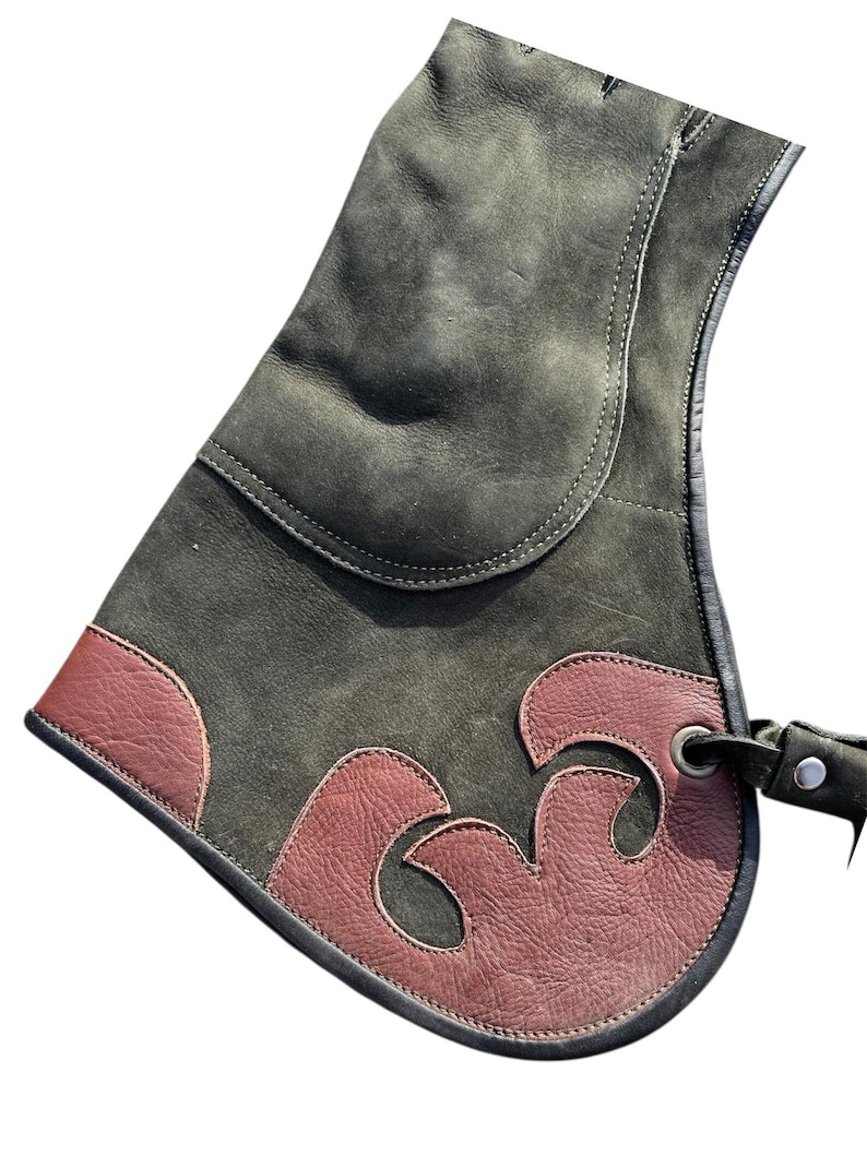 May include: A dark green leather falconry glove with a decorative brown leather accent. The glove features a curved design and a strap with a metal eyelet for securing it to the wrist. The glove is designed to protect the wearer's hand.