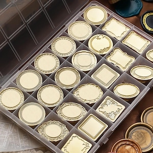 Può includere: Un organizer in plastica trasparente pieno di vari timbri per sigilli di cera color oro. I timbri hanno forme diverse, tra cui cerchi, ovali e rettangoli, con bordi e disegni decorativi. I timbri sono disposti in scomparti individuali.
