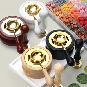 May include: An assortment of wax seal stamp warmers and wax seal stamps. The warmers are made of wood, black, and white materials with gold-colored metal accents. The wax seal stamps have wooden and black handles. A container of colorful wax beads is also visible.