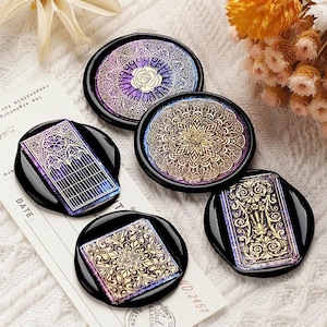 May include: A collection of five black, round, and rectangular decorative wax seals with intricate gold and iridescent designs. The seals feature floral, geometric, and architectural patterns. The background includes a white surface and a piece of paper with text.