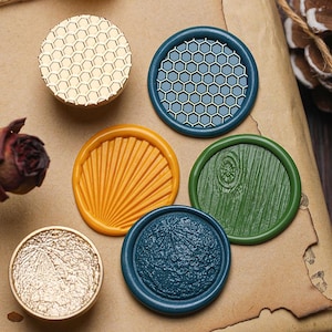 May include: A selection of decorative wax seals in various colours and designs. The seals feature gold, blue, green, and orange hues, with patterns including honeycomb, wood grain, and radial lines. These seals are used for adding a decorative touch to letters and crafts.