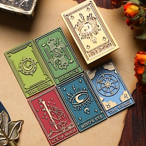 May include: A collection of tarot card-themed pins in various colors, including green, red, blue, and gold. Each pin features intricate designs and text, such as "THE GUIDE," "BALANCE," "ACE OF WANDS," "MIND'S EYE," and "THE WHEEL."