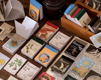 Könnte beinhalten: Eine Sammlung von Papieren im Vintage-Stil, darunter kleine Notizbücher mit farbenfrohen Einbänden, Postkarten und dekoratives Papier. Die Notizbücher zeigen Illustrationen und Texte in verschiedenen Schriftarten und Farben, mit einem Fokus auf botanische und himmlische Themen.
