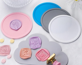 May include: A collection of craft supplies including a pink wax seal stamp with a gold handle, a gray cloud-shaped mat with wax seals, and several colorful silicone coasters. The wax seals feature floral and botanical designs in various colors.