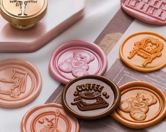 May include: A collection of decorative wax seals in various colors and designs. Seals include a teddy bear with "I Love You", a coffee cup, a chair, and a bicycle. A gold-colored lamp-shaped stamp is also visible. The seals are arranged on a patterned surface.