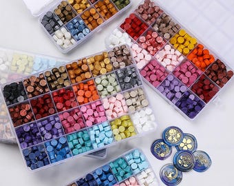 May include: Assortment of colorful wax seal beads in clear plastic organizer boxes. The beads are in various shapes and shades, including red, blue, green, and purple. Several decorative wax seals with gold accents are also visible.