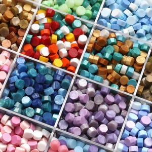 Könnte beinhalten: Eine Vielzahl farbenfroher Wachssiegelperlen in einem transparenten, unterteilten Behälter. Die Perlen sind in Rot-, Orange-, Gelb-, Weiß-, Blau-, Grün- und Lilatönen gehalten. Die sechseckigen Perlen sind für Bastelarbeiten und Schreibwaren.
