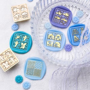 Pode incluir: Uma coleção de selos de cera decorativos em várias cores, incluindo dourado, verde-azulado e roxo. Cada selo apresenta um design único, como anjos, padrões climáticos e ursos. Os selos estão dispostos em um prato de vidro transparente, com pequenos enfeites em forma de flor.