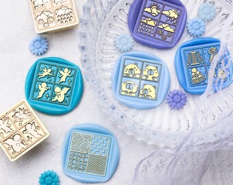 May include: A collection of decorative wax seals in various colors, including gold, teal, and purple. Each seal features a unique design, such as angels, weather patterns, and bears. The seals are arranged on a clear glass dish, with small flower-shaped embellishments.