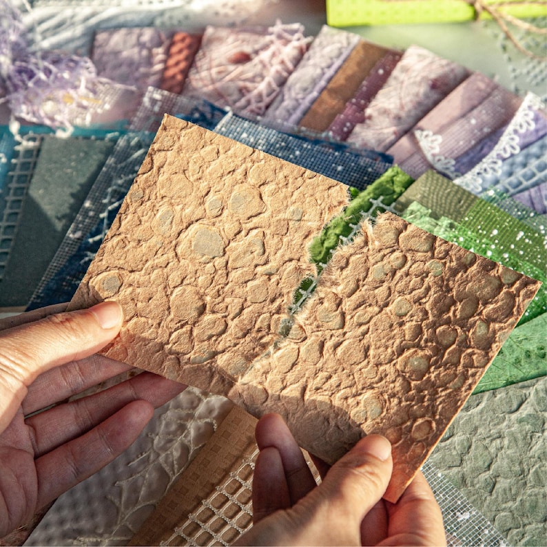 May include: A person holding a textured, light brown paper square with a pebble-like pattern. Various other patterned papers in shades of blue, green, and purple are visible in the background, suggesting a craft or scrapbooking theme.