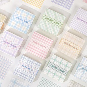 May include: A collection of small, square boxes with plaid patterns in pastel colors. Each box has a label with text such as "SEASALT SODA," "CHESTNUT CAKE," and "FLESHY GRAPE." The boxes are arranged on a white surface.