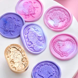 May include: A collection of colorful wax seals in shades of pink, purple, and gold. The seals feature various designs, including floral bouquets, heart-shaped motifs, and whimsical characters. The gold seal has a rectangular shape.