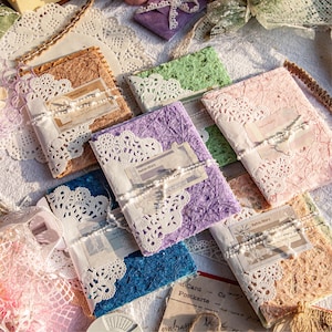 May include: A collection of handmade journals in various colors, including brown, green, purple, pink, and blue. Each journal is adorned with white lace and tied with a ribbon. The journals have a vintage aesthetic, with the text "Retro Story" visible on some.