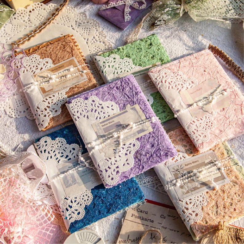 May include: A collection of handmade journals in various colors, including brown, green, purple, pink, and blue. Each journal is adorned with white lace and tied with a ribbon. The journals have a vintage aesthetic, with the text "Retro Story" visible on some.