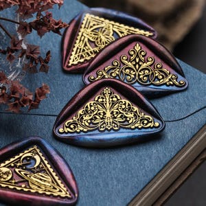 Może przedstawiać: Zbliżenie kilku dekoracyjnych pieczęci woskowych ze złotymi wzorami filigranowymi. Pieczęcie są trójkątne z gradientem fioletu, niebieskiego i złota. Jedna pieczęć ma motyw motyla, podczas gdy inne mają bogate wzory kwiatowe i motywy niebieskie.