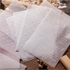 Peut inclure: Une collection de feuilles de papier calque translucide présentant des motifs en relief, notamment des cœurs, des fleurs, des papillons, des lapins, des étoiles, des lunes et du texte en écriture cursive. Papier semi-transparent texturé.