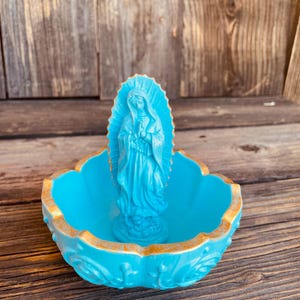 May include: A turquoise Virgin Mary statue stands in a matching decorative bowl with a gold rim. The bowl has a scalloped edge and ornate detailing. The statue is centered in the bowl, creating a religious or spiritual decorative piece.