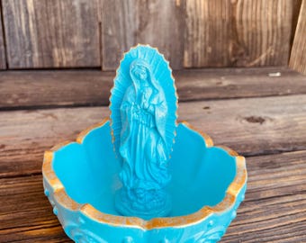Puede incluir: Una estatua turquesa de la Virgen María se encuentra en un cuenco decorativo a juego con un borde dorado. El cuenco tiene un borde festoneado y detalles ornamentados. La estatua está centrada en el cuenco, creando una pieza decorativa religiosa o espiritual.