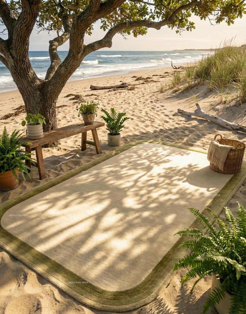 K&ouml;nnte beinhalten: Ein rechteckiger beiger Teppich mit gr&uuml;nem Rand liegt an einem Sandstrand. Eine Holzbank mit Topfpflanzen steht unter einem gro&szlig;en Baum. Ein Weidenkorb mit einer Decke und weiteren Pflanzen befindet sich in der N&auml;he. Das Meer ist im Hintergrund zu sehen.