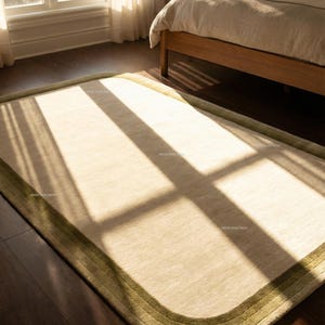 K&ouml;nnte beinhalten: Beiger rechteckiger Teppich mit abgerundeten Ecken und gr&uuml;nem Rand. Der Teppich liegt auf einem dunklen Holzboden in einem Schlafzimmer, mit Sonnenlicht, das durch ein Fenster scheint. Ungef&auml;hre Gr&ouml;&szlig;e: 200 cm x 300 cm.