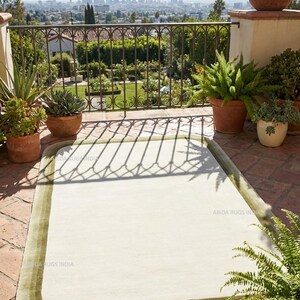 K&ouml;nnte beinhalten: Ein rechteckiger, cremefarbener Teppich mit gr&uuml;nem Rand liegt auf einer Backstein-Terrasse. Die Aufnahme zeigt Topfpflanzen und ein schmiedeeisernes Gel&auml;nder. Im Hintergrund ist eine Stadtlandschaft zu sehen.