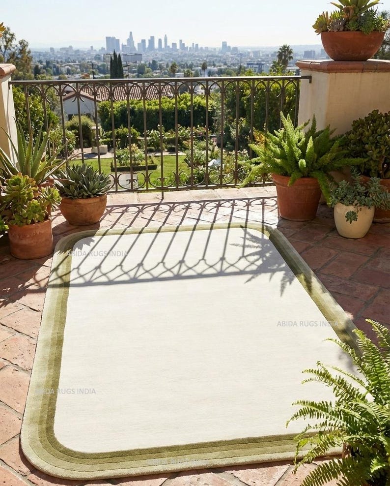 K&ouml;nnte beinhalten: Ein rechteckiger, cremefarbener Teppich mit gr&uuml;nem Rand liegt auf einer Backstein-Terrasse. Die Aufnahme zeigt Topfpflanzen und ein schmiedeeisernes Gel&auml;nder. Im Hintergrund ist eine Stadtlandschaft zu sehen.