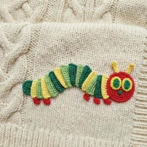Puede incluir: Una aplicación de oruga de ganchillo en tonos verdes, amarillos y rojos, con una cara sonriente y pequeños pies rojos, está cosida sobre una tela de punto de cable color crema. La oruga es un diseño caprichoso.
