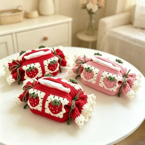 Puede incluir: Tres fundas de caja de pañuelos hechas a mano con ganchillo en forma de caramelos. Dos son rojas con detalles blancos y verdes, y una es rosa con detalles blancos y verdes. Cada una presenta un diseño de fresa y bordes con volantes.