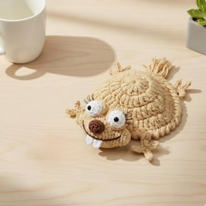 Può includere: Un sottobicchiere all'uncinetto beige a forma di lucertola dei cartoni animati, con occhi bianchi, ciglia, naso marrone e due denti bianchi. Il sottobicchiere è su una superficie di legno chiaro.