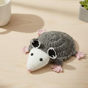 May include: A handmade crochet opossum coaster in gray, white, black, and pink. The coaster features a round body, a white snout, black eyes, and pink feet. The opossum is lying down on a light wooden surface.