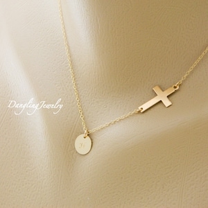 May include: A gold necklace with a small round pendant and a delicate cross charm. The pendant has a small initial engraved on it.