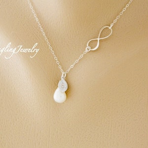 May include: A silver necklace with an infinity symbol charm and a pearl pendant. The pearl is attached to a small silver disc with a letter engraved on it.