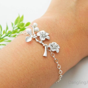 May include: A silver bracelet with a delicate branch design featuring three blossoms and pearl accents. The bracelet is on a person's wrist.