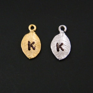 May include: Two leaf-shaped charms with the letter "K" engraved on them. One charm is gold and the other is silver.