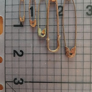 Può includere: Una collezione di cinque spille di sicurezza vintage di varie dimensioni, disposte verticalmente. Le spille sono in metallo arrugginito e invecchiato, con usura visibile. Sono poste su uno sfondo a griglia grigia, con i numeri 1, 2 e 3 visibili.