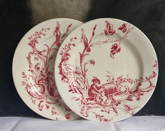 Puede incluir: Dos platos color crema con un patrón de toile rojo. Los platos presentan un diseño detallado de figuras, elementos florales y volutas decorativas. Los platos tienen una estética clásica y vintage.