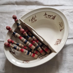 Könnte beinhalten: Eine ovale, geteilte Servierschale mit cremefarbener Glasur und braunen Illustrationen. Die Schale enthält mehrere Holzpuppen mit roten, blauen und naturfarbenen Holzstreifen. Die Puppen haben rote, kugelförmige Köpfe.