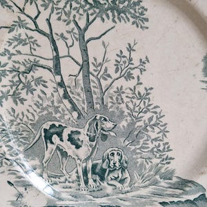 May include: Off-white ceramic plate with a teal illustration of two dogs in a forest scene. One dog is standing, the other is lying down. Trees and foliage surround the dogs, with a body of water in the foreground.